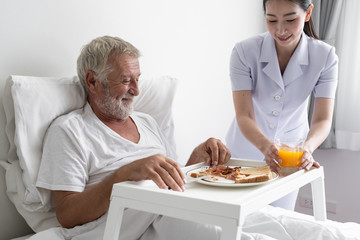 senior man with smiling nurse, takes care breakfast and discussion and cheer on bed at nursing home