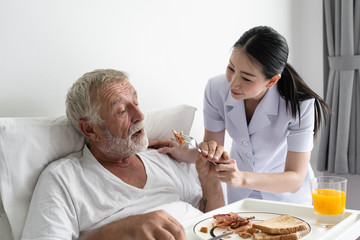 senior man with smiling nurse, takes care breakfast and discussion and cheer on bed at nursing home