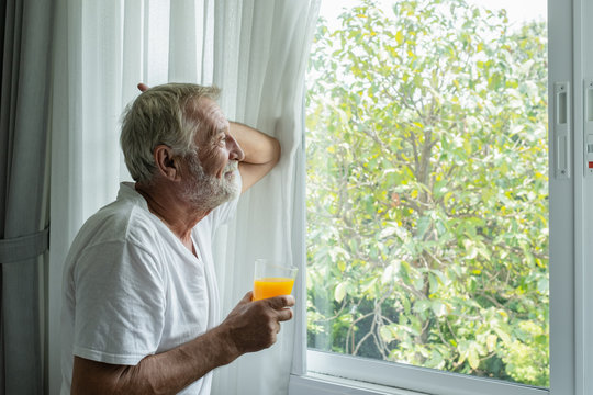 senior man hold glass of orange juice and thinking and looking outside of window in white room