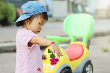 In the summer season. Happy Asian baby child girl holding pink flowers in her hand and playing a car toy at the playground. Baby aged of one year old and 4 months. Expression of kid concept.