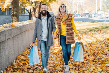 Autumn fun. Couple walking with shopping bags along sidewalk covered with yellow leaves. Fall city landscape background.