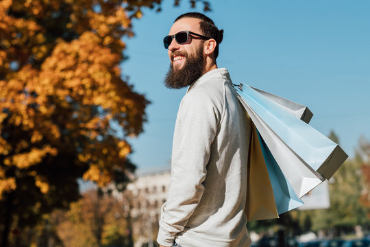 Man fashion style. Bearded hipster guy with shopping bags enjoying autumn sunny day. Blur fall trees and blue sky background.