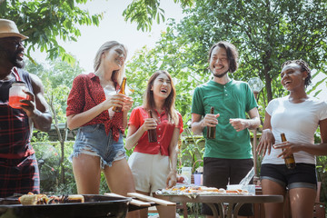 Group of happy friends standing eating and drinking beers at barbecue dinner camping in nature and having meal together outdoor as summer lifestyle, food and friendship concept