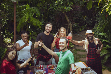 Group of happy friends standing eating and drinking beers at barbecue dinner camping in nature and having meal together outdoor as summer lifestyle, food and friendship concept