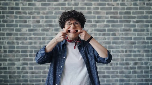 Portrait of playful young woman making funny faces and smiling having fun looking at camera standing against brick wall background. Cheerful people and joy concept.