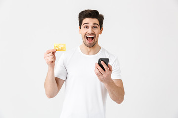 Excited happy young man posing isolated over white wall background using mobile phone holding credit card.