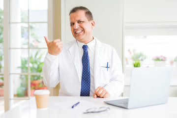 Middle age doctor man wearing white medical coat working with laptop at the clinic smiling with happy face looking and pointing to the side with thumb up.