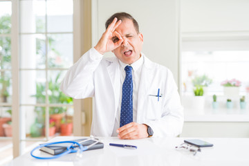 Middle age doctor man wearing medical coat at the clinic doing ok gesture with hand smiling, eye looking through fingers with happy face.