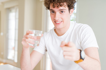 Young man drinking a glass of water at home pointing with finger to the camera and to you, hand sign, positive and confident gesture from the front