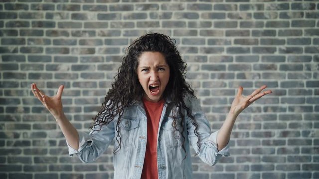 Portrait of mad young woman hipster screaming with anger standing on brick background raising arms shouting looking at camera. People and emotions concept.