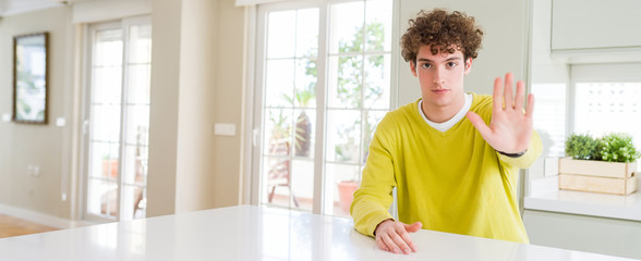 Wide angle shot of young handsome man at home doing stop sing with palm of the hand. Warning expression with negative and serious gesture on the face.