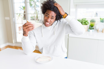 Fototapeta na wymiar African American man eating handmade sandwich at home stressed with hand on head, shocked with shame and surprise face, angry and frustrated. Fear and upset for mistake.