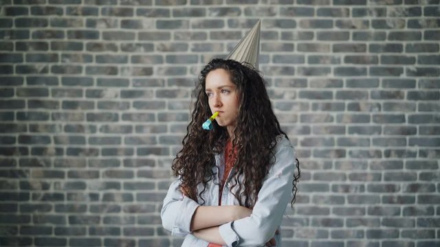 Portrait of upset birthday girl in party hat blowing whistle standing with crossed arms and sad face on brick background. Bad mood, disappointing holidays and sadness concept.