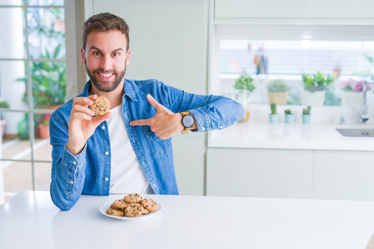 Handsome man eating chocolate chips cookies with surprise face pointing finger to himself