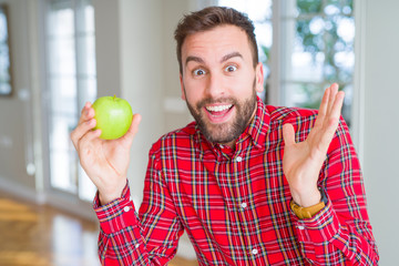 Handsome man eating fresh healthy green apple very happy and excited, winner expression celebrating victory screaming with big smile and raised hands