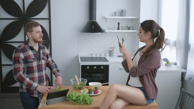cheerful girl sitting on table and taking pictures funny guy on cell phone while cooking useful meal for brunch in kitchen