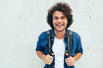 Outdoors horizontal portrait of young man with curly hair, smiling broadly, with backpack on the back, standing at building concrete background on the city street. People and lifestyle. - Powered by Adobe