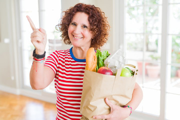 Senior woman holding paper bag full of fresh groceries from the supermarket very happy pointing with hand and finger to the side