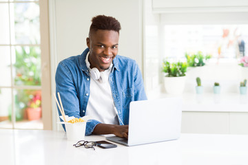 Handsome young african business man eating delivery asian food and working using computer, enjoying noodles smiling