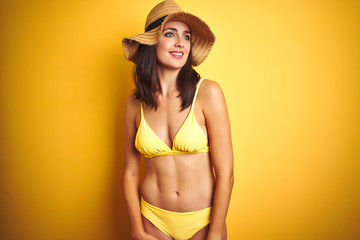 Beautiful woman wearing yellow bikini and summer hat over isolated yellow background looking away to side with smile on face, natural expression. Laughing confident.
