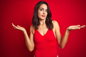 Beautiful woman wearing bikini swimwear over red isolated background clueless and confused expression with arms and hands raised. Doubt concept.