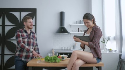 diet plan, happy guy prepares delicious healthy salad from fresh vegetables and greens and girl sits on table with tablet in arms and talks in kitchen