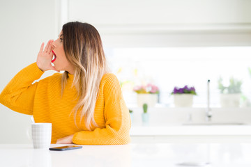 Young beautiful woman drinking a cup of coffee at home shouting and screaming loud to side with hand on mouth. Communication concept.