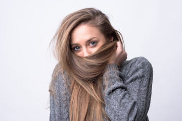 Portrait of playful young woman holding long hair as scarf hiding and covering on light gray background. 