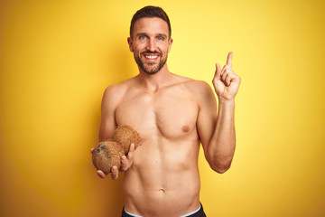 Fototapeta premium Young handsome shirtless man holding exotic tropical coconut over isolated yellow background very happy pointing with hand and finger to the side
