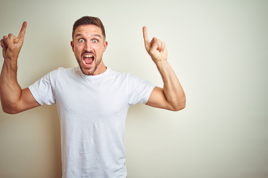 Young handsome man wearing casual white t-shirt over isolated background smiling amazed and surprised and pointing up with fingers and raised arms.