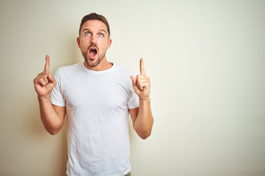 Young handsome man wearing casual white t-shirt over isolated background amazed and surprised looking up and pointing with fingers and raised arms.