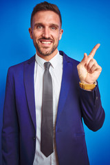 Vertical angle picture of young handsome business man over blue isolated background with a big smile on face, pointing with hand and finger to the side looking at the camera.