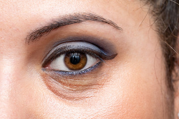 A closeup view on the open eye of a beautiful young Caucasian woman, showing blue eyeshadow with dark puffy circles beneath the eyes, a common symptom of sleep deprivation.