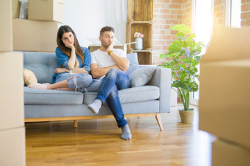 Young beautiful couple relaxing sitting on the sofa around boxes from moving to new house thinking looking tired and bored with depression problems with crossed arms.