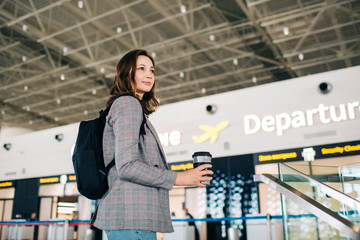 Young attractive traveler girl with backpack and coffee cup turning around at the departure zone at the airport. Travel vibes.