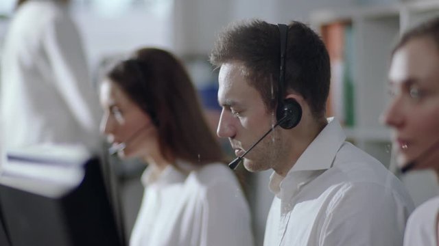 Side view on working operators, selective focus on positive handsome Caucasian male customer services agent with headset working in a call center, smiling and talking to customer