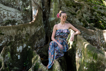 a girl in a blue dress sits in the roots of a banyan tree. The tree is old and overgrown with moss