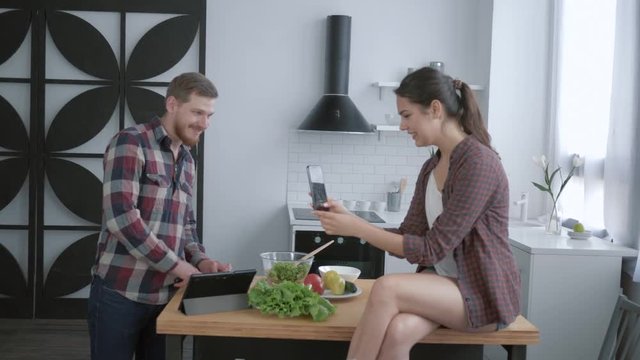 funny merry guy with girl are photographed on mobile phone while cooking healthy food for dinner and make grimaces of vegetables in bright kitchen