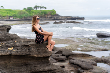 Fototapeta na wymiar girl in a dress sits on the rocky shore of the ocean and looks into the distance, enjoying the calm water