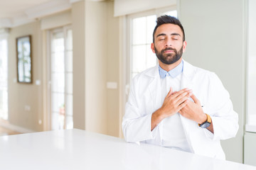 Fototapeta premium Handsome hispanic doctor or therapist man wearing medical coat at the clinic smiling with hands on chest with closed eyes and grateful gesture on face. Health concept.