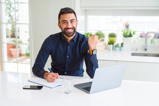 Handsome hispanic man working using computer and writing on a paper pointing and showing with thumb up to the side with happy face smiling