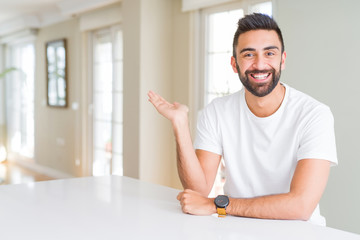 Handsome hispanic man casual white t-shirt at home smiling cheerful presenting and pointing with palm of hand looking at the camera.