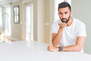 Fototapeta premium Handsome hispanic man casual white t-shirt at home looking at the camera blowing a kiss with hand on air being lovely and sexy. Love expression.