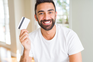 Handsome hispanic man holding credit card with a happy face standing and smiling with a confident...