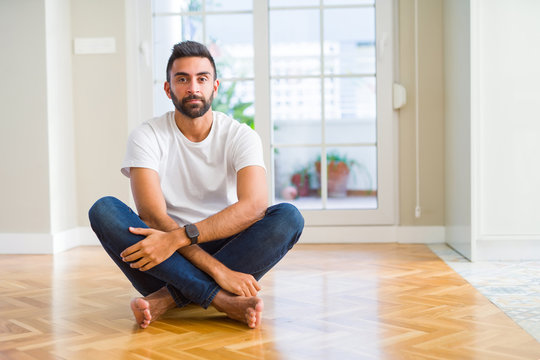 Handsome hispanic man wearing casual t-shirt sitting on the floor at home with serious expression on face. Simple and natural looking at the camera.