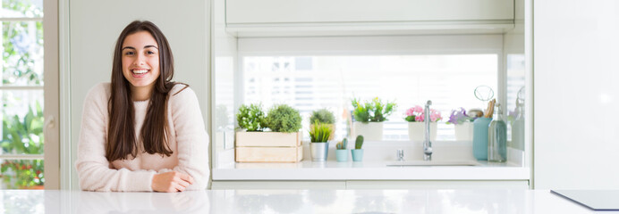 Wide angle picture of beautiful young woman sitting on white table at home with a happy and cool smile on face. Lucky person.