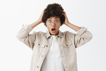 Studio shot of impressed and surprised emotive african american in glasses and beige boyish shirt holding hand on head gasping and smiling broadly being amazed and astonished of recent news