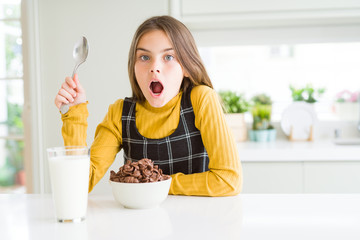 Beautiful young girl kid eating chocolate cereals and glass of milk for breakfast scared in shock...