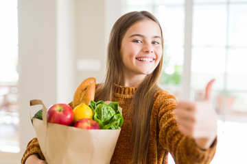 Beautiful young girl holding paper bag of fresh groceries happy with big smile doing ok sign, thumb up with fingers, excellent sign