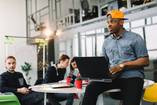 Dark skinned African american coworker in jeans shirt and yellow cap and using laptop in modern office. Multiracial young creative people are working together with laptop, tablet, smart phone.